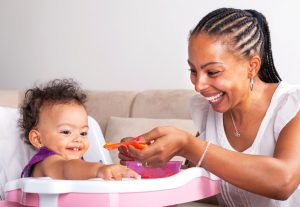 African-American Mother Feeding Baby Girl In High Chair