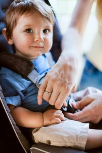 A mom loads her cute infant son safely into a rear facing car seat as they get ready to drive somewhere in their vehicle. The baby smiles at the camera. Sunlight shines in from behind. Vertical.