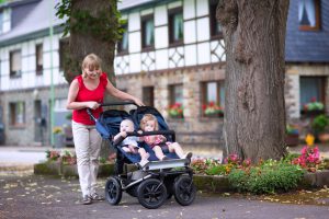 Young beautiful woman with a double jogging stroller with two kids, baby boy and little toddler girl, brother and sister, walking in a beautiful German village with traditional old houses