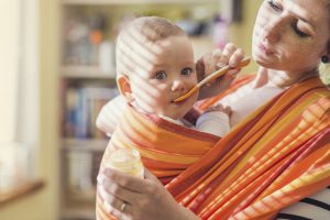 Young mother feeding her little daughter that she has in a baby carrier