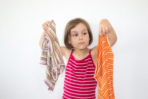 unstable little girl looking at clothings to select one. Little girl raising two casual clothings and looking at them to choose. She is confused. Can not decide. Standing in front of wall of home. Horizontal composition. Image developed from Raw.