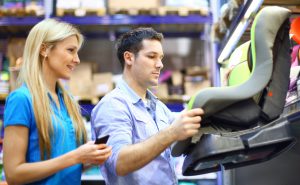 Closeup side view of early 30's couple choosing a baby seat to install in their car. The man is holding the seat and reading the specs. His wife is just checking the visual side of the seat. Both are wearing casual clothes, she's in blue polo t-shirt and he's wearing regular light blue shirt with rolled up sleeves.
