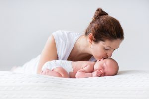Young mother hugging her newborn child. Mom nursing baby. Woman and new born boy relax in a white bedroom. Family at home. Love, trust and tenderness concept. Bedding and textile for nursery.