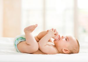 Cute baby boy lying down in bed and putting his feet into mouth.