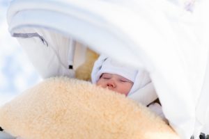Cute newborn child sleeping in stroller on a cold winter day. New born baby taking a nap in warm sheepskin fur footmuff in a stroller in snowy park. Kids sleep outdoors in pram. Family fun in snow.